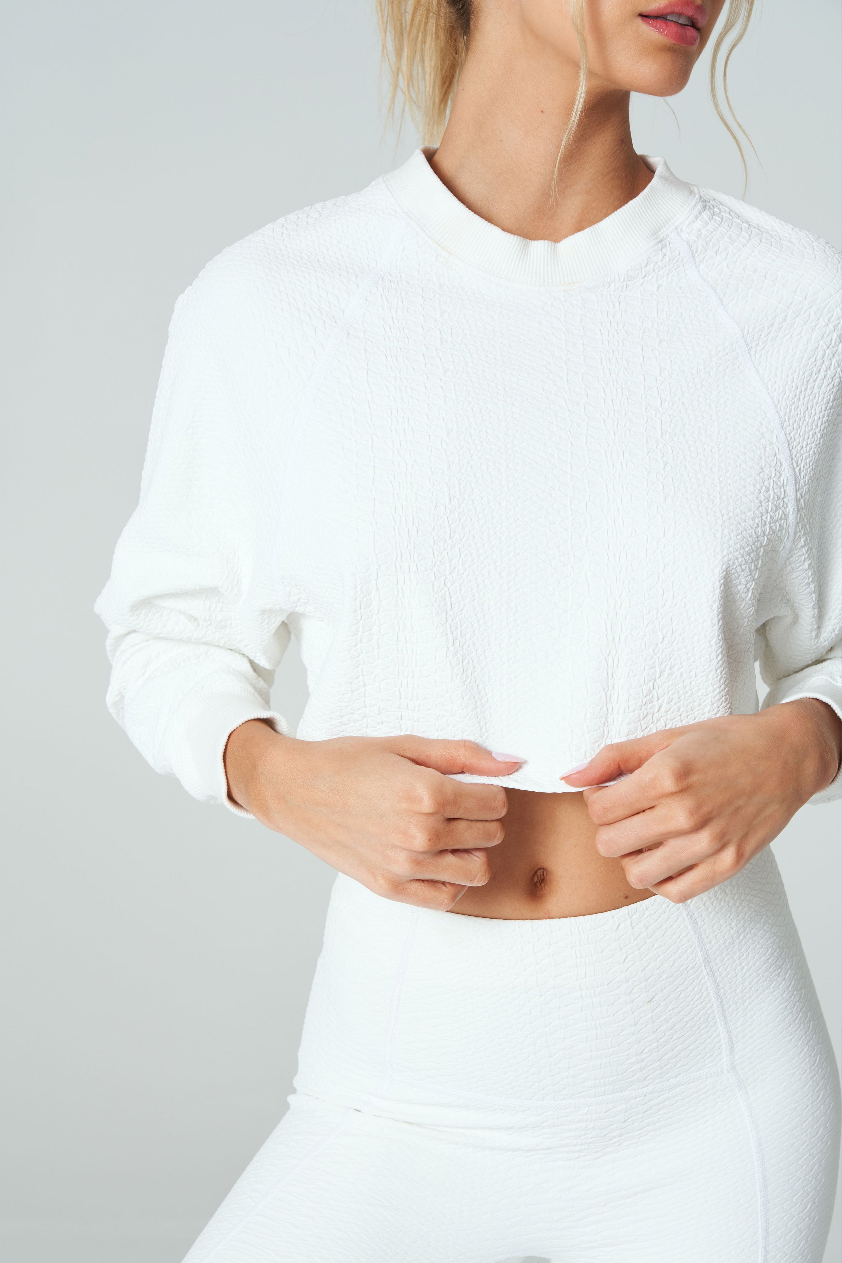 A woman in the Isabella Pullover by Gigi C Sport and matching pants stands against a light gray background. Her blonde hair is tied back, and she is partially cropped out from the shoulders up, adding an air of mystery.