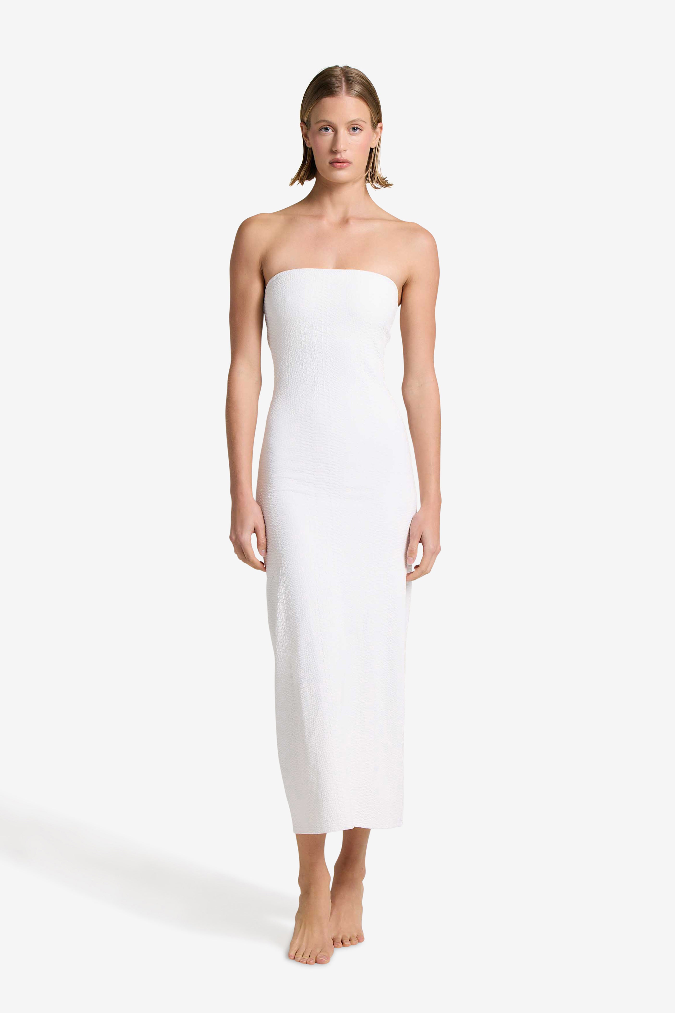 Barefoot against a plain backdrop, a person wears Gigi C Bikinis elegant Maya Dress in ivory. The strapless, ankle-length design complements their relaxed posture as short, straight hair frames their serene demeanor, with arms at their sides.