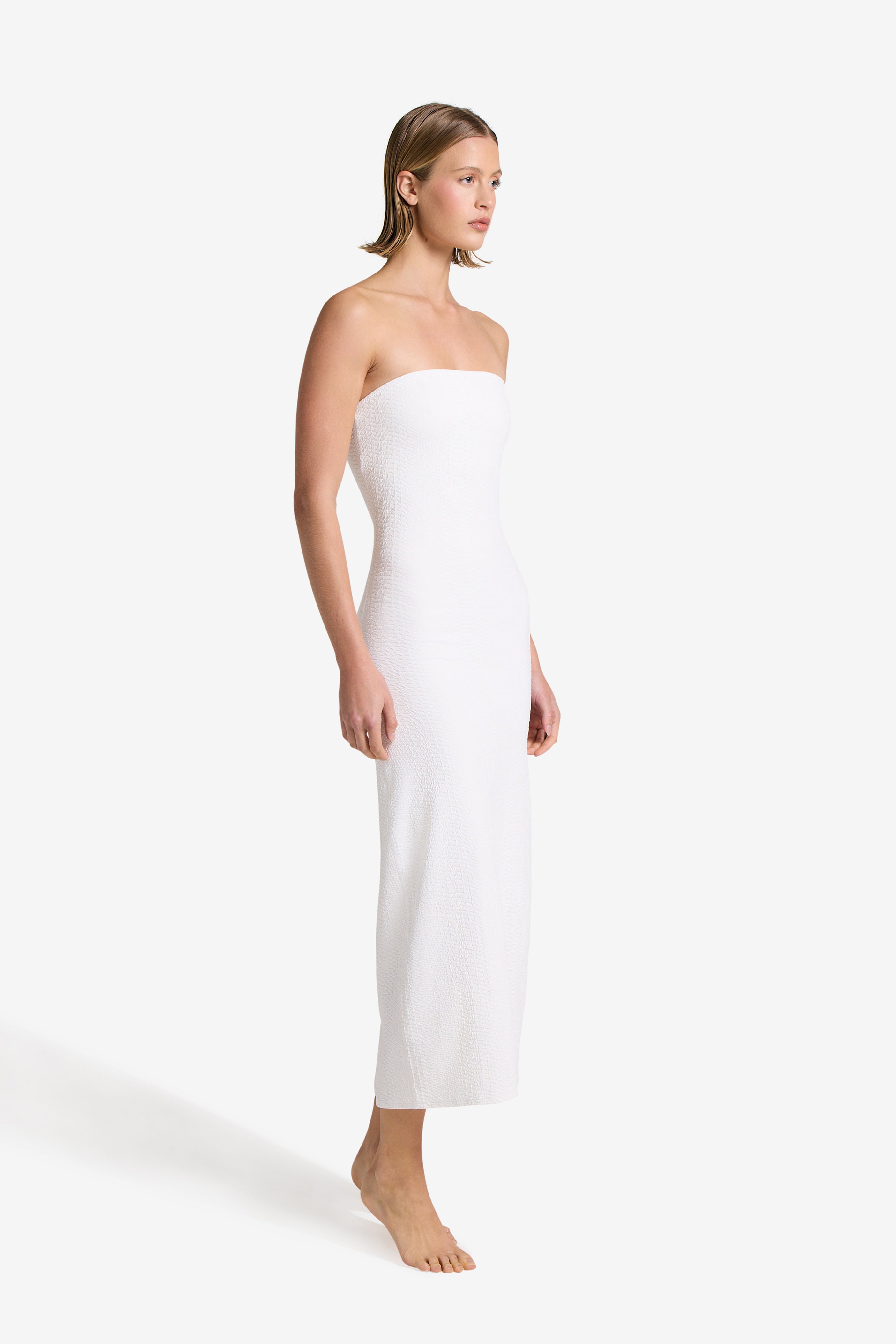 A woman stands barefoot against a light background, wearing an ivory ankle-length Maya Dress by Gigi C Bikinis. Facing slightly left with relaxed arms, her short, neatly styled hair enhances an elegant look.