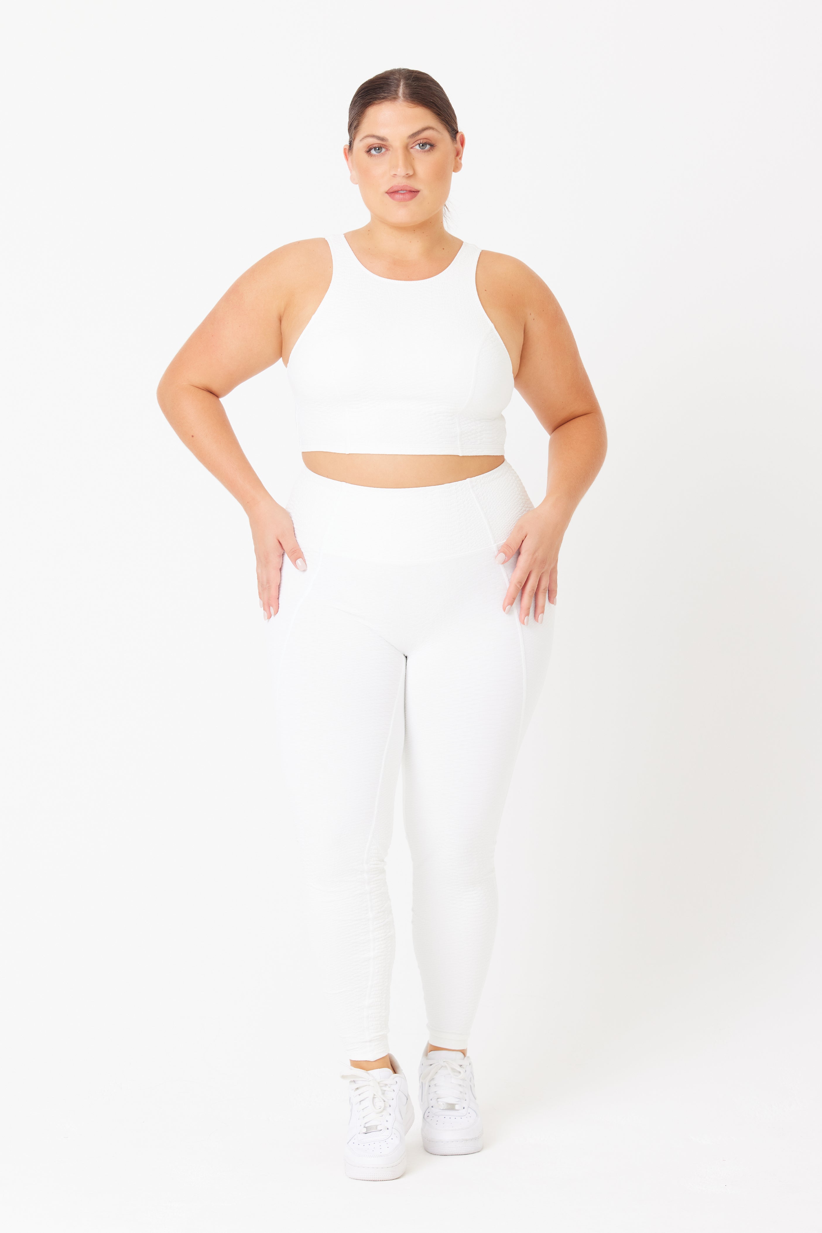 Against a white background, a person wears Gigi C Sports Ava Legging and a white sports bra. They pair it with faux snakeskin sneakers. With their hair tied back, they gaze directly at the camera with a neutral expression.