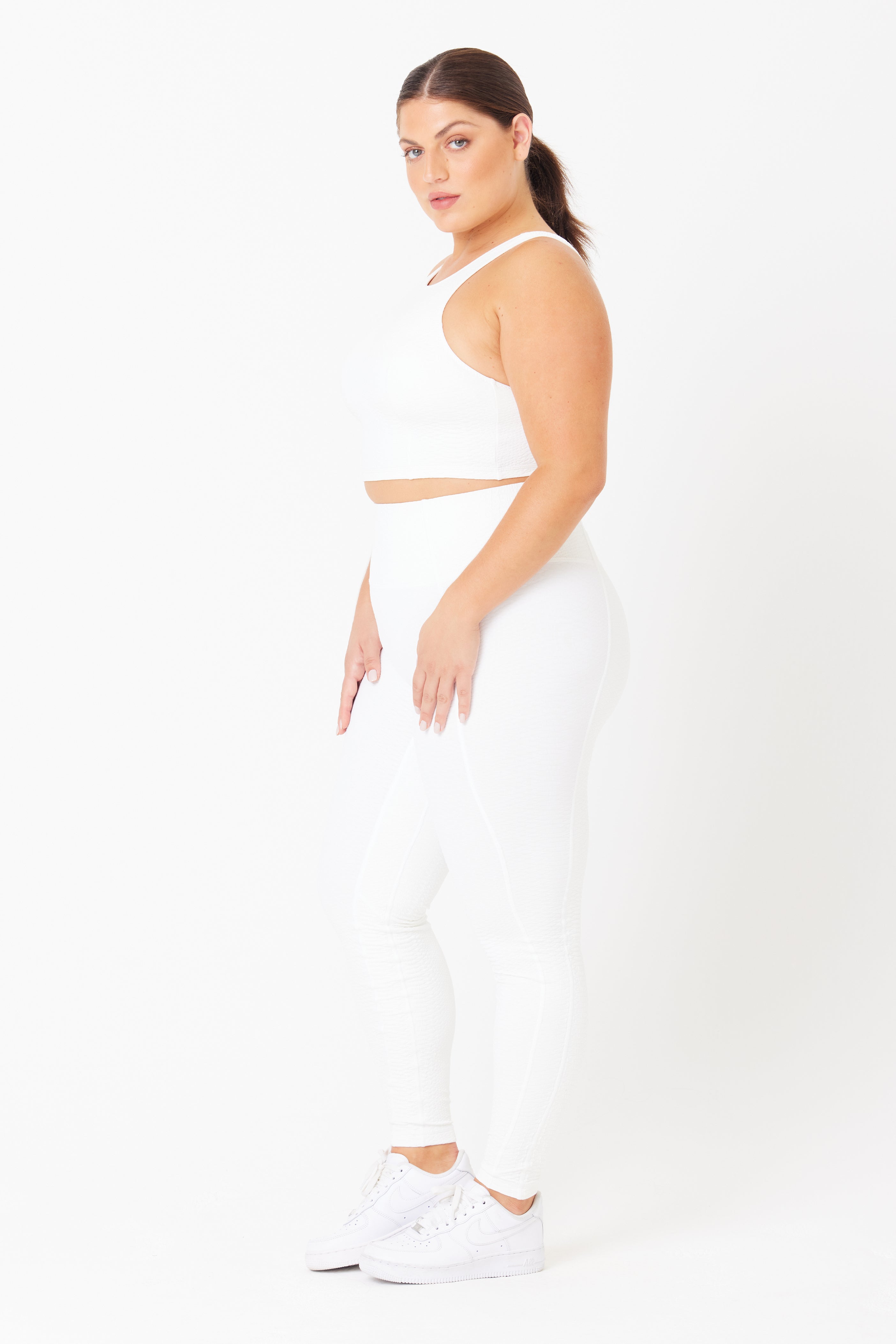 A model in a Gigi C Sport white sports bra, Ava Legging with bi-stretch compression, and matching white sneakers stands sideways on a white background. They have dark hair tied back and maintain a neutral expression, facing slightly toward the camera.