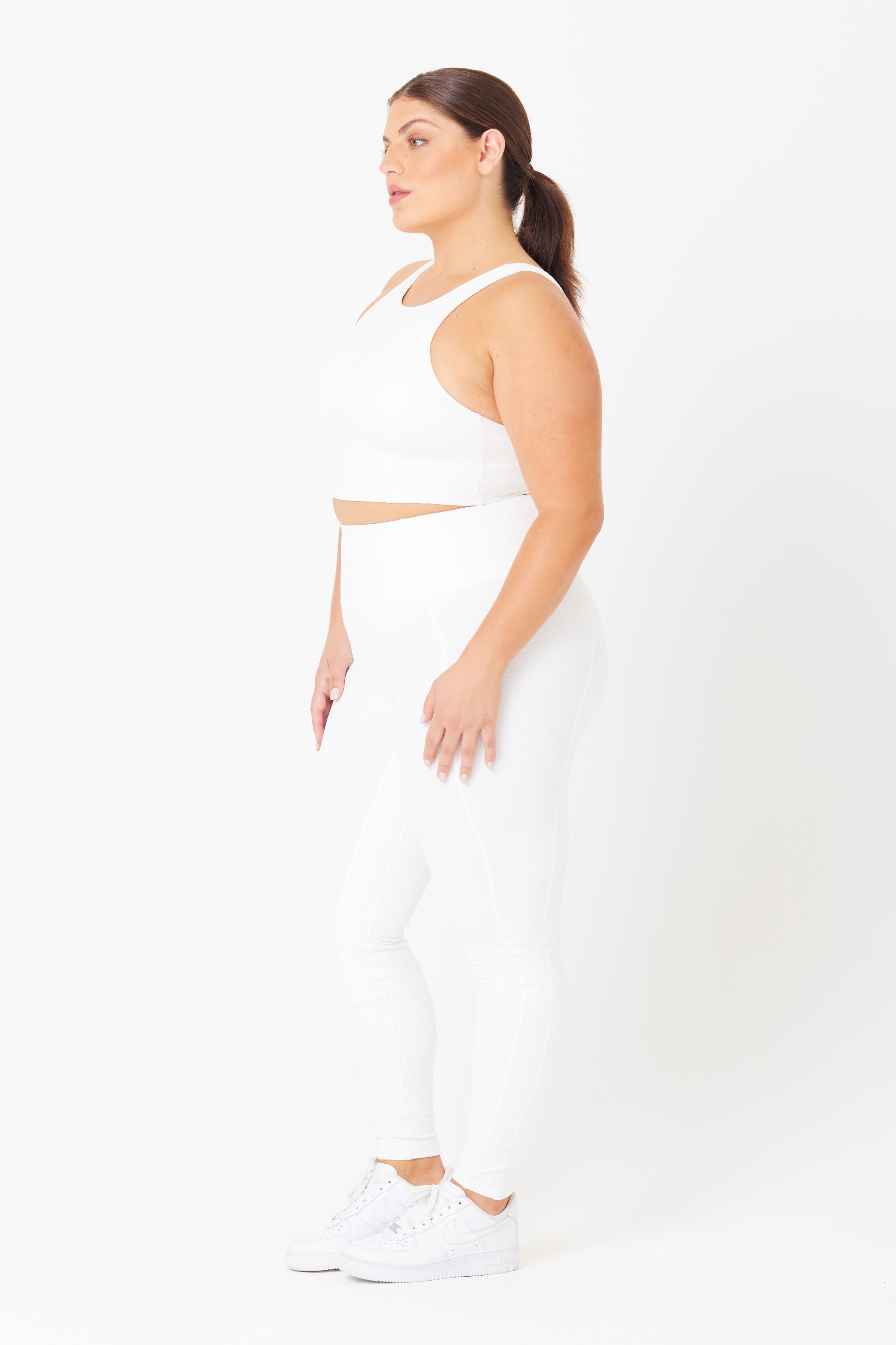A person is shown in profile on a white background, wearing a white athletic crop top, high-waist Ava Legging by Gigi C Sport in bi-stretch compression fabric, and white sneakers. Their hair is styled in a ponytail.