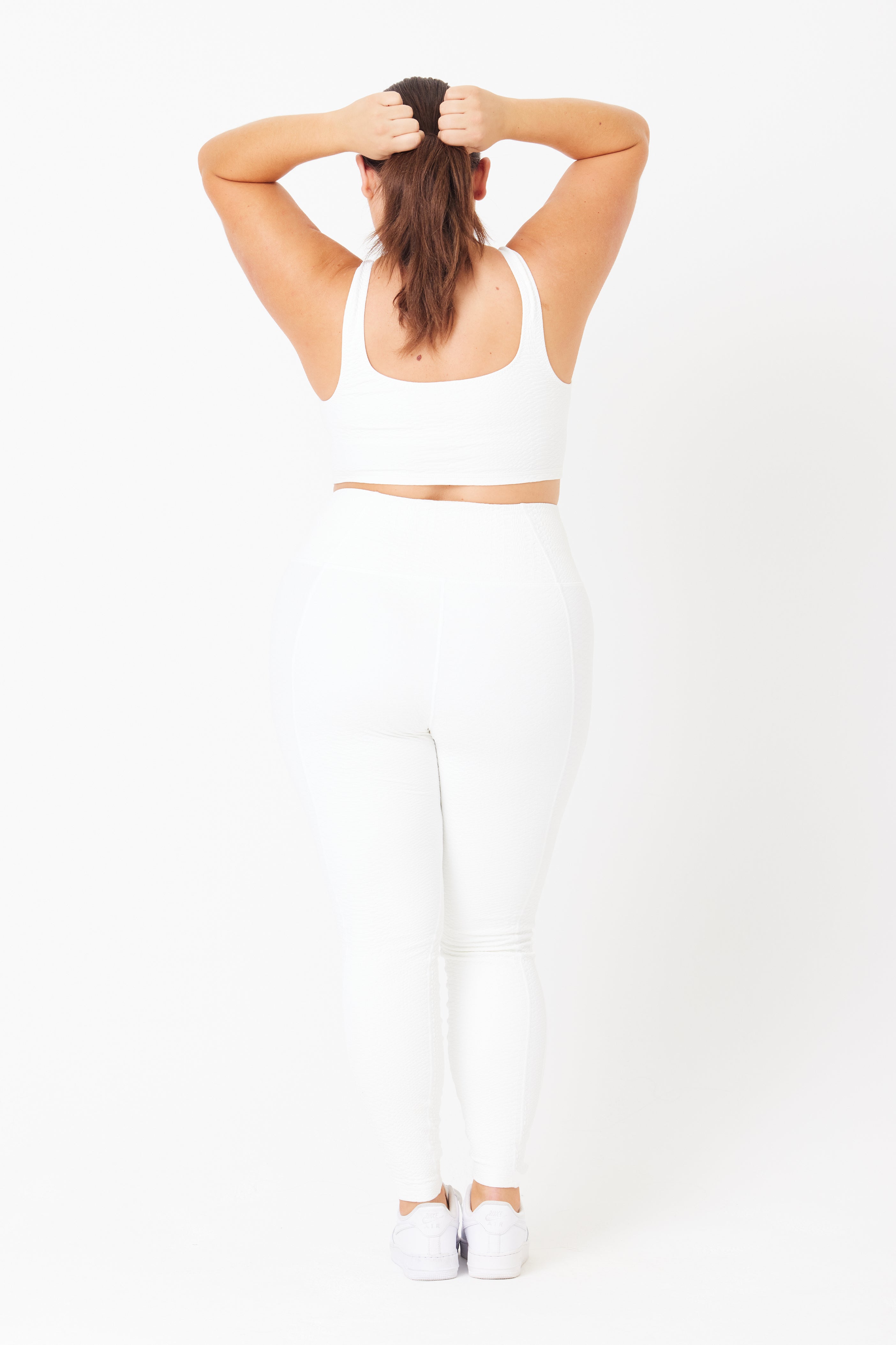 A person with long hair stands facing away, holding their hair up while wearing a white sports bra and the high-waist Ava Legging featuring bi-stretch compression from Gigi C Sport. Set against a plain white background, they complete the look with matching sneakers.