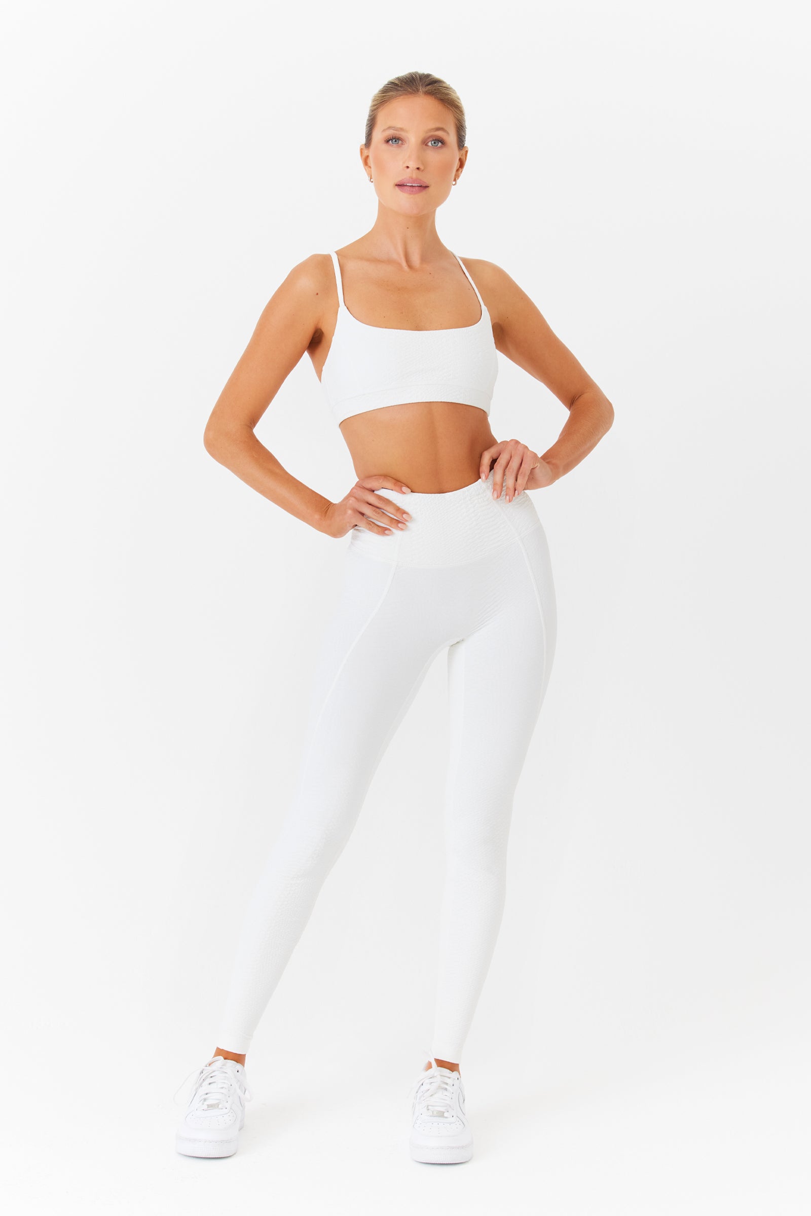 A person in a white athletic outfit poses confidently against a plain white background, wearing a sports bra, Gigi C Sports Ava Legging with bi-stretch compression, and white sneakers.