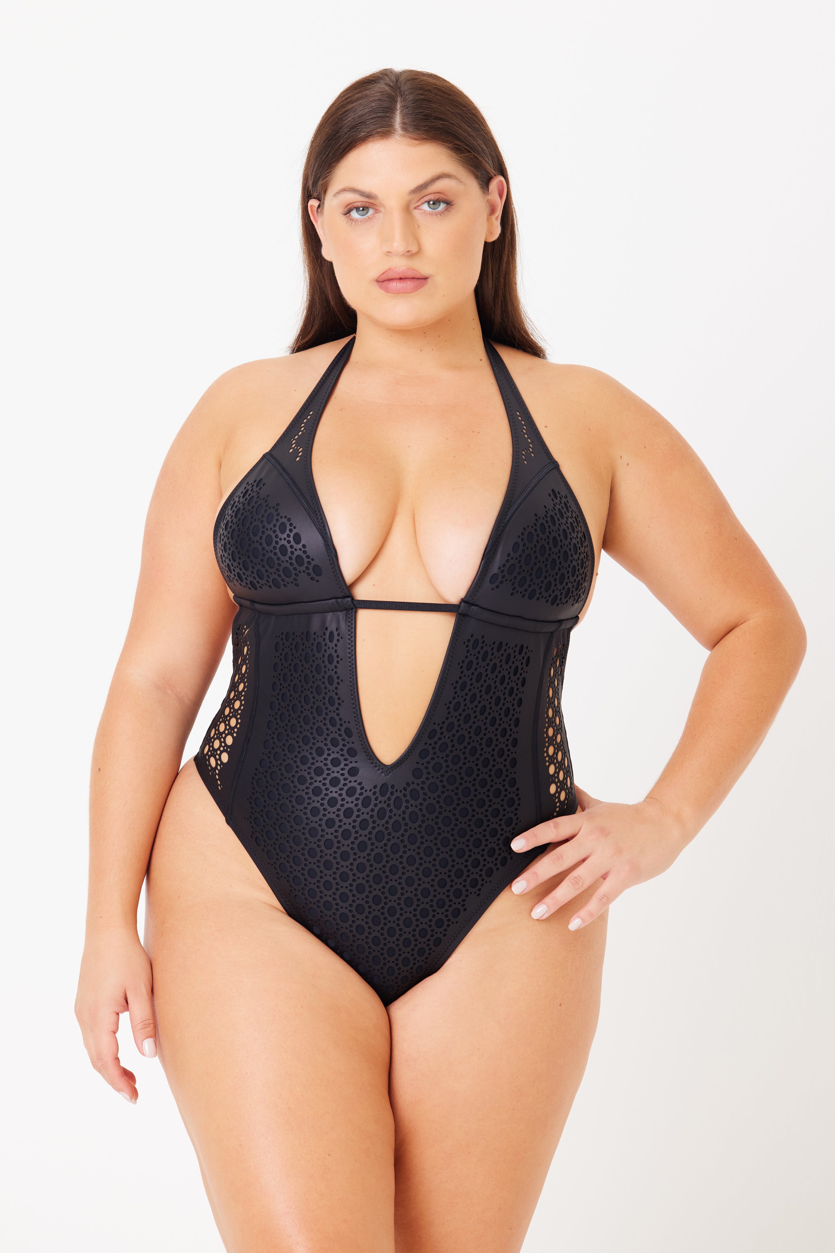 A model showcases the Camilla One Piece by Gigi C Bikinis, a black swimsuit featuring intricate laser cutouts and a geometric design. It has removable bust pads, a deep V-neckline, and comfortable halter straps, all set against a stark white background.