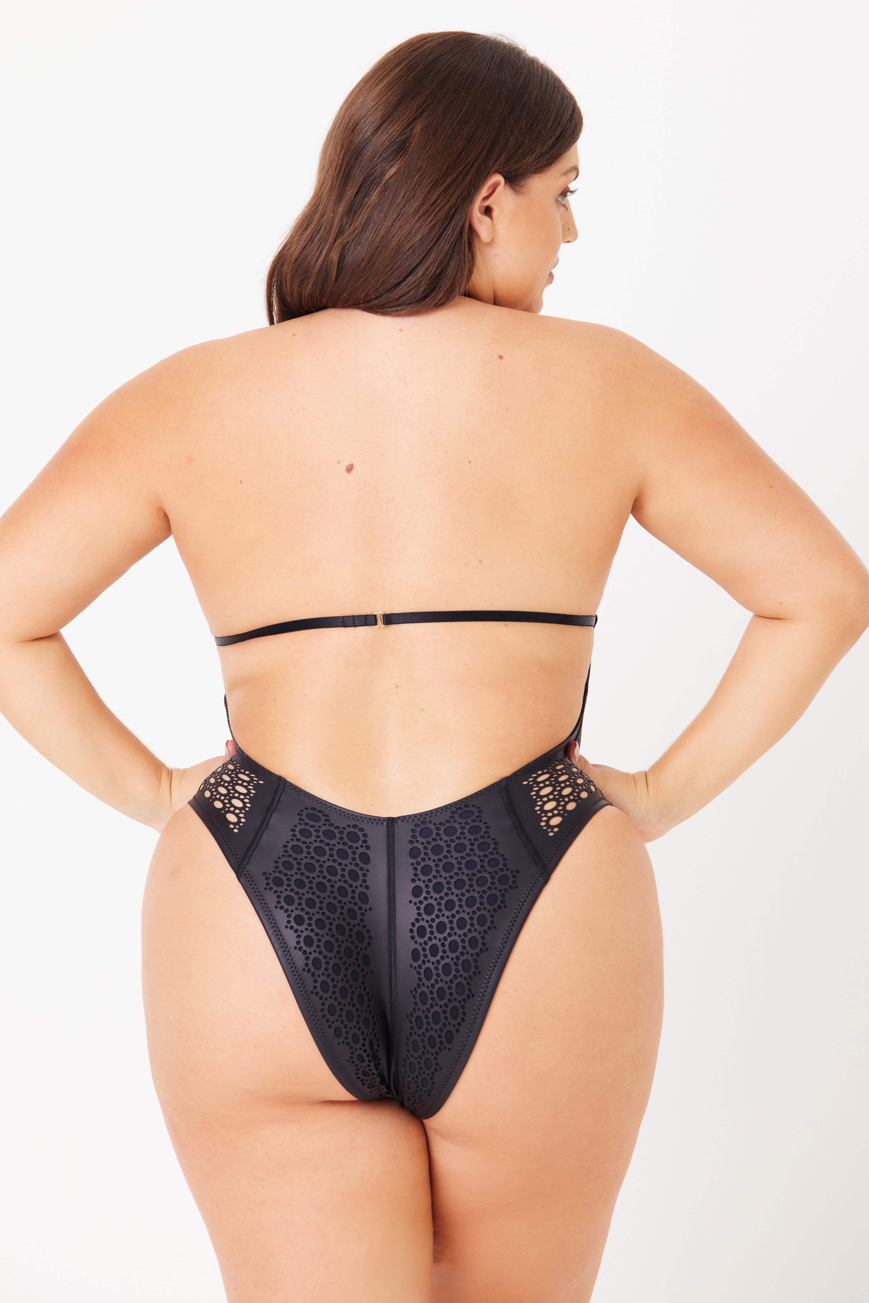 A person is seen from the rear wearing the Gigi C Bikinis Camilla One Piece—a high-cut black lace bodysuit with back strap detailing and intricate laser cutouts, offering minimal coverage against a plain white background.
