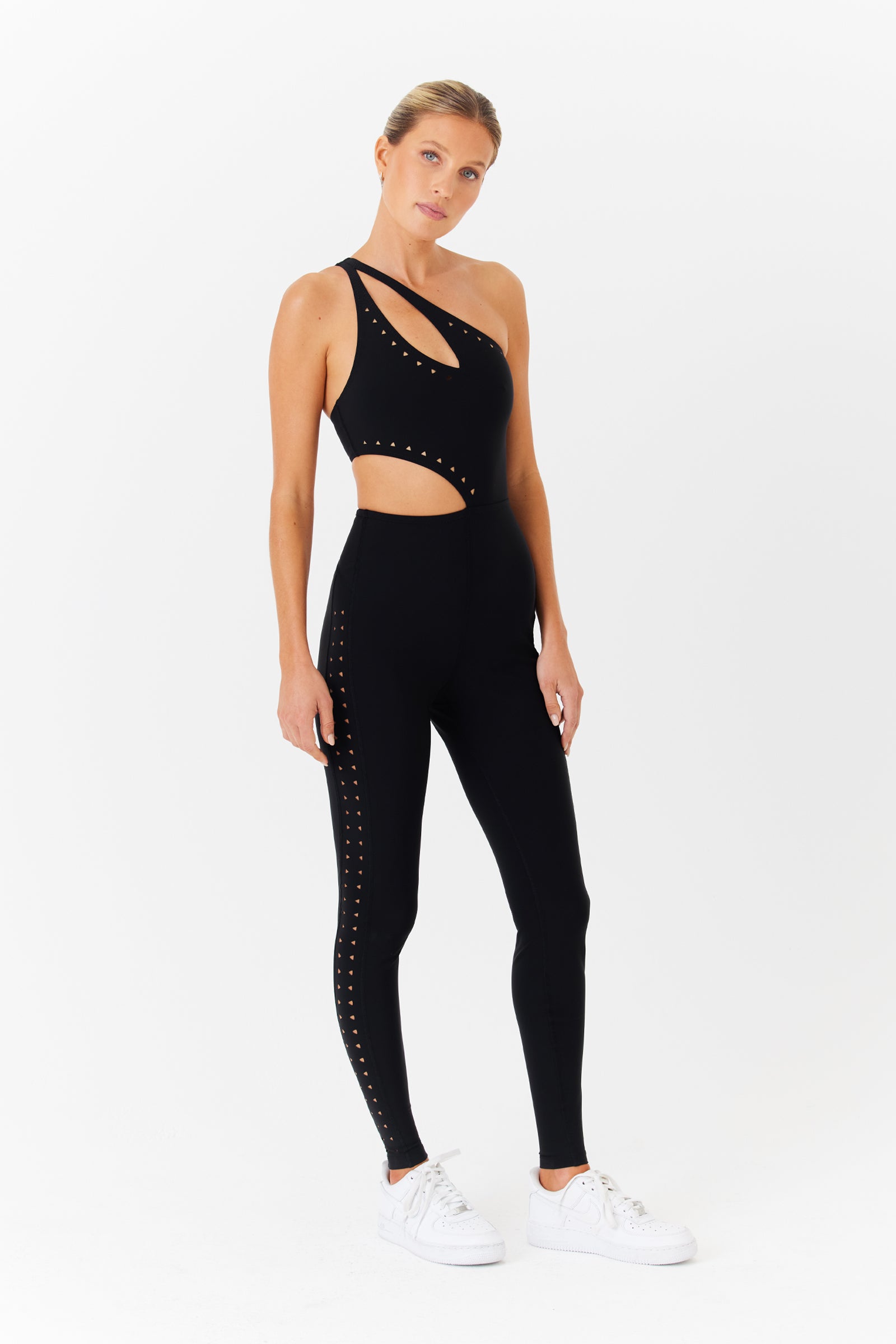 A person stands against a white background wearing a black Cassie Jumpsuit by Gigi C Sport, featuring an asymmetrical design, intricate laser cutouts, and decorative stud patterns. The breathable knit ensures comfort. White sneakers complete the look.