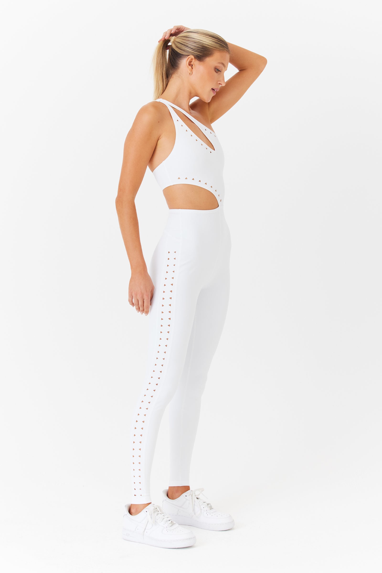 The woman poses in profile wearing a stunning Gigi C Sport Cassie Jumpsuit with laser cutouts and studded details against a white background. Her sleeveless outfit in breathable knit pairs effortlessly with white sneakers as she casually raises a hand to her ponytail-adorned head.