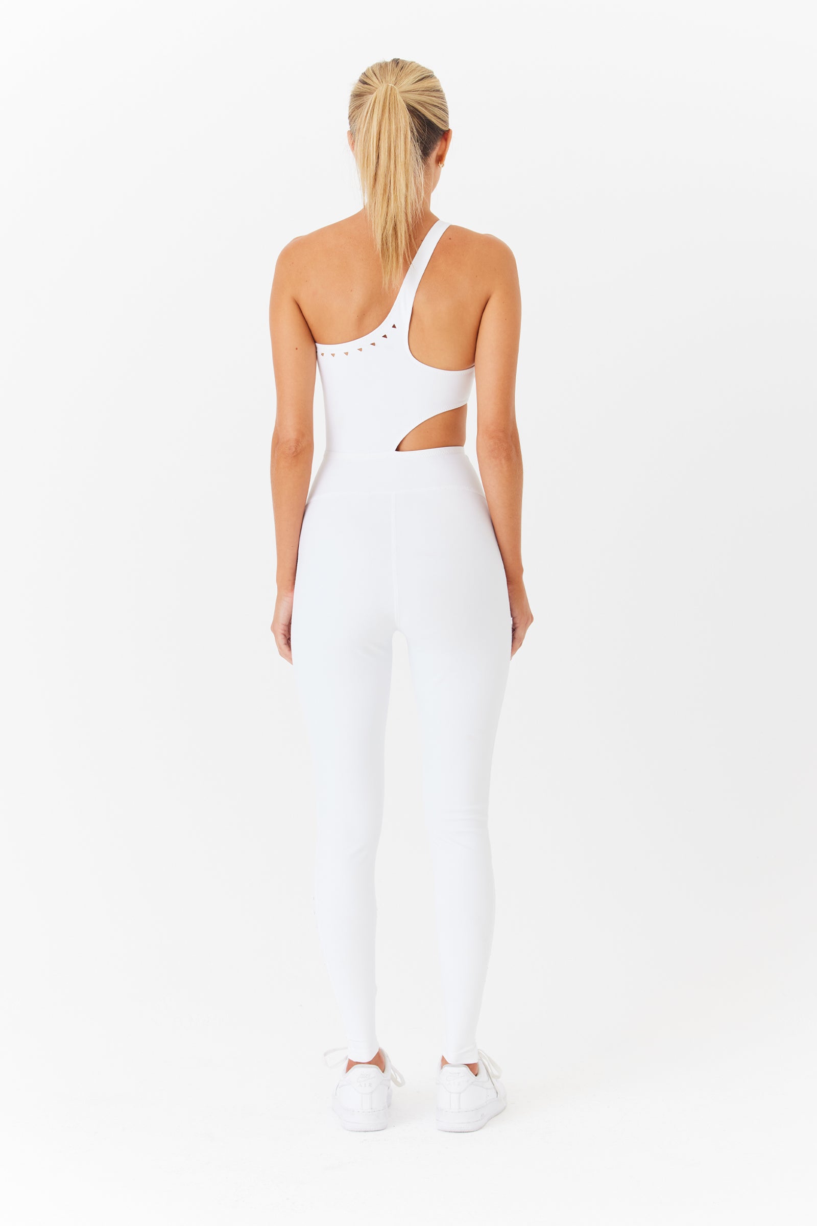 A person with long blonde hair stands facing away, wearing a Gigi C Sport Cassie jumpsuit with laser cutouts and an asymmetric design enhanced by shoulder and side details. They effortlessly pair it with white sneakers against a plain white backdrop.