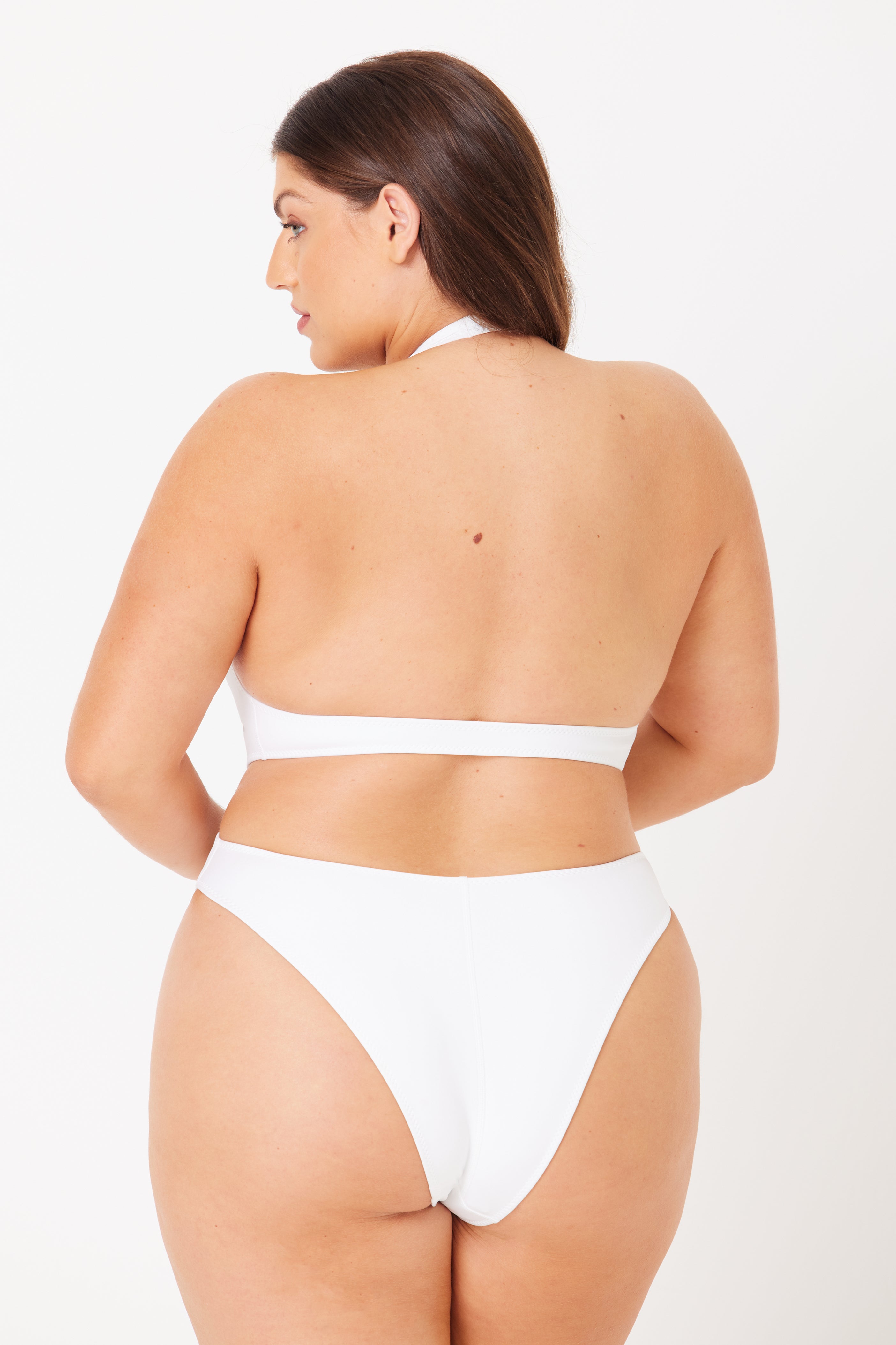 A person with long, dark hair, seen from the back, wears the Charlie Plunging One-Piece by Gigi C Bikinis. The white halter swimsuit provides cheeky coverage as they stand against a plain white background with their head turned slightly to the right.