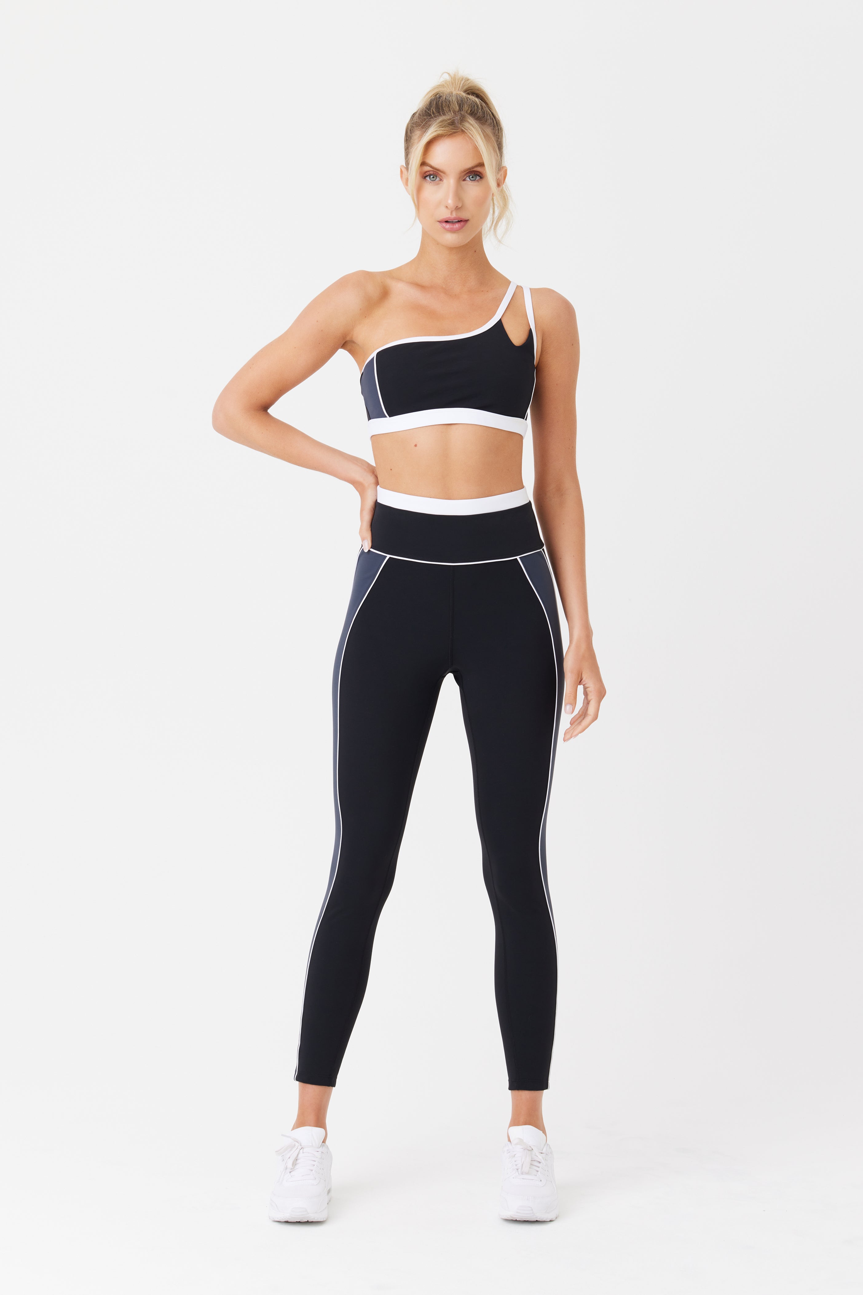 A woman models a black and white sporty look with Gigi C Sports Faith Asymmetrical Bra and high-waisted leggings against a white backdrop. She pairs it with white sneakers and an updo, capturing hot yoga vibes.