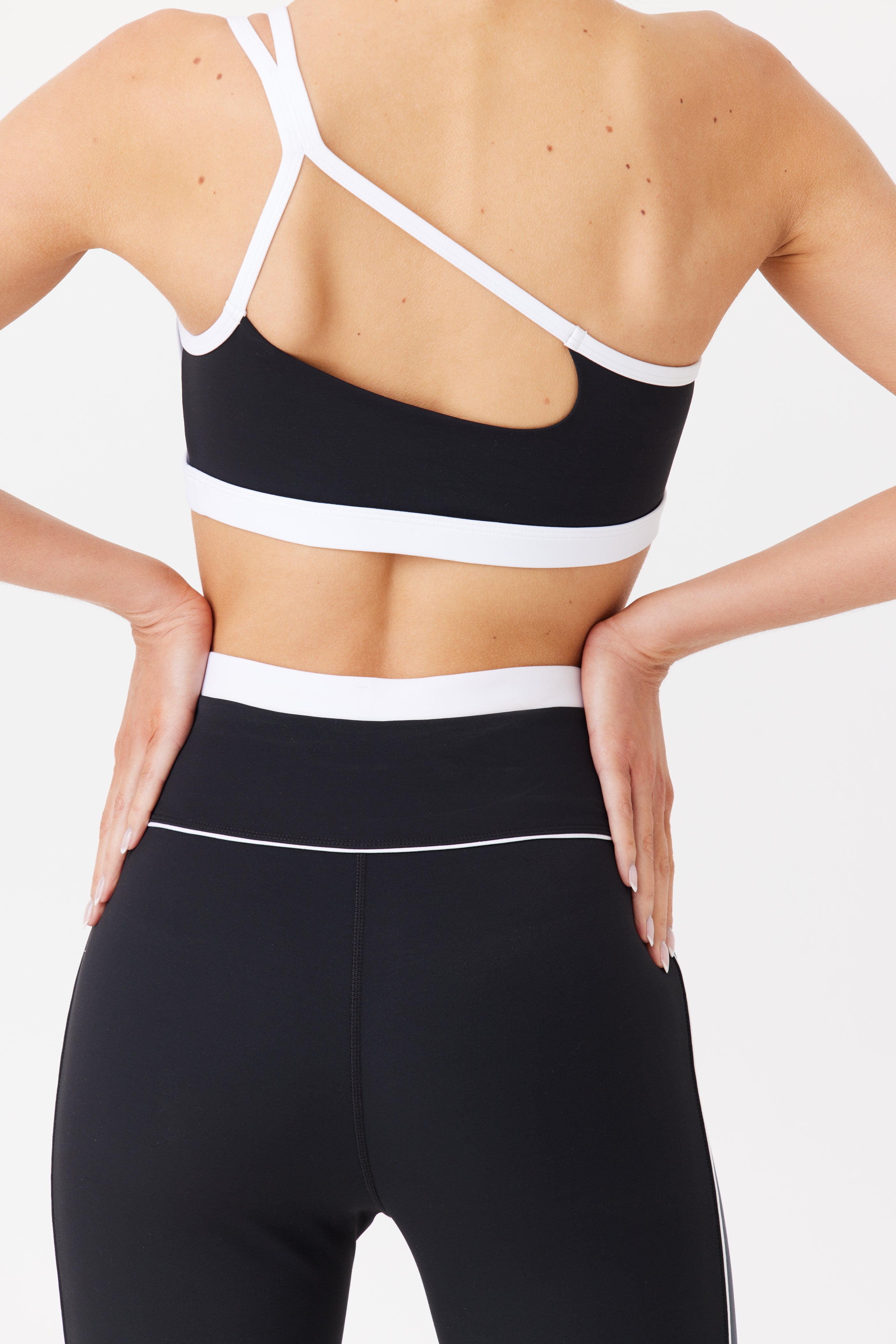 A person wears the Faith Asymmetrical Bra by Gigi C Sport with white straps and high-waisted leggings, showcasing a cutout design from behind. Ideal for hot yoga, they stand confidently hands on hips against a plain white background.