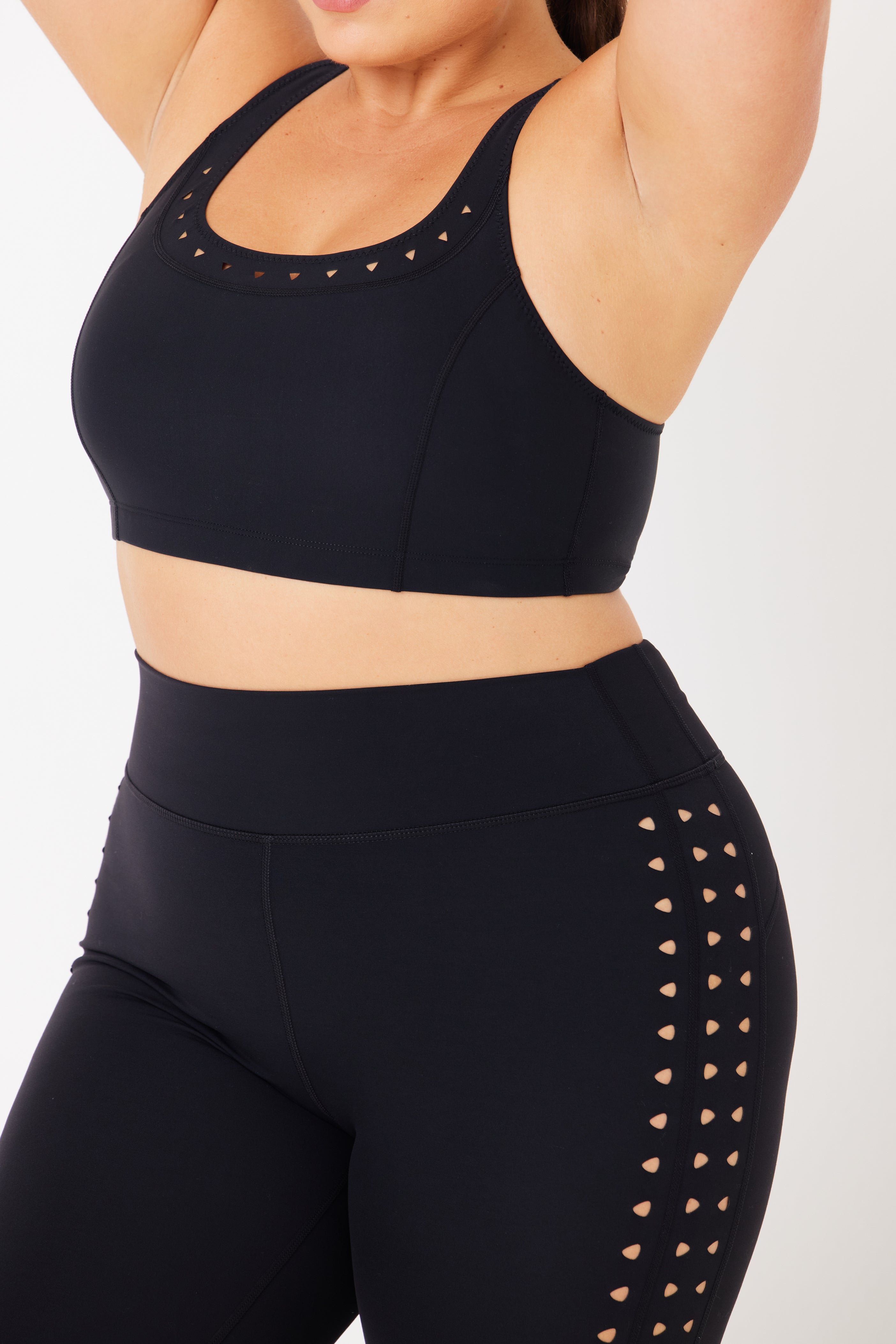 A person raises their arms wearing a black Gigi C Sport sports bra and Katrina Legging, featuring compression fabric with laser cutouts on the neckline and sides, set against a plain white background.