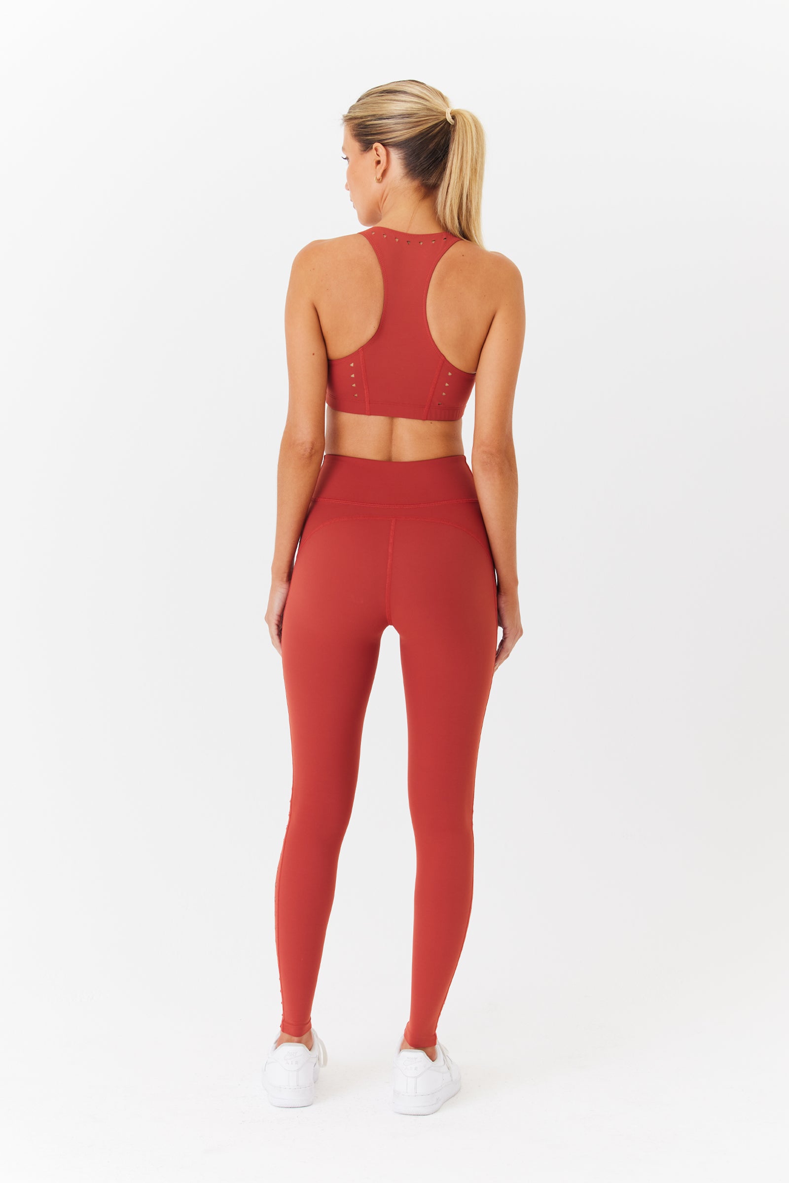 A woman with a ponytail faces away, wearing Gigi C Sports Annabelle Bra Top and leggings in a red, super-soft fabric. Shes paired them with white sneakers against a plain white background.