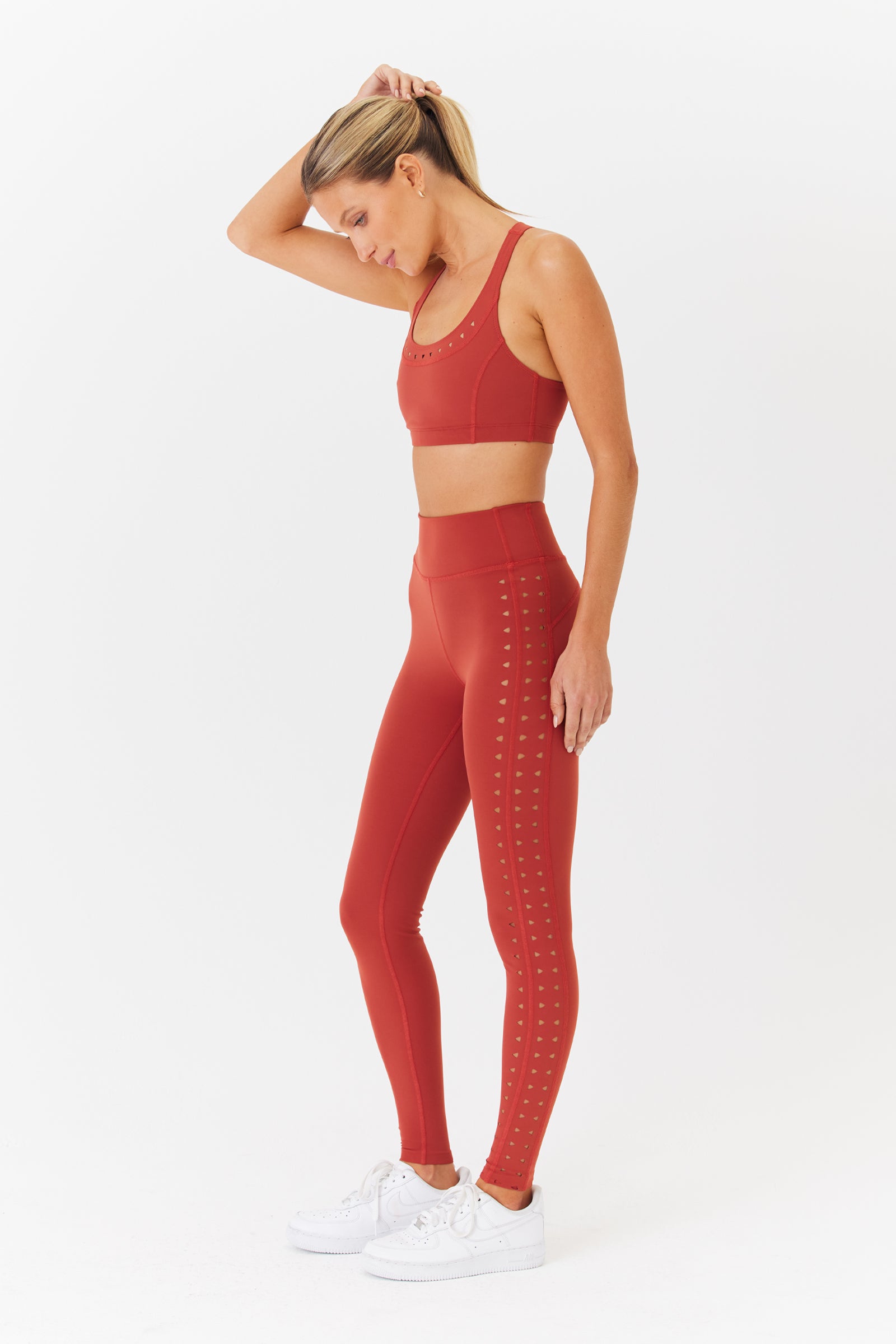 A woman in profile against a white background wears Gigi C Sport Katrina Legging and a red sports bra, both made from compression fabric that contours her curves. She completes her look with white sneakers and gently pulls back her hair with one hand.