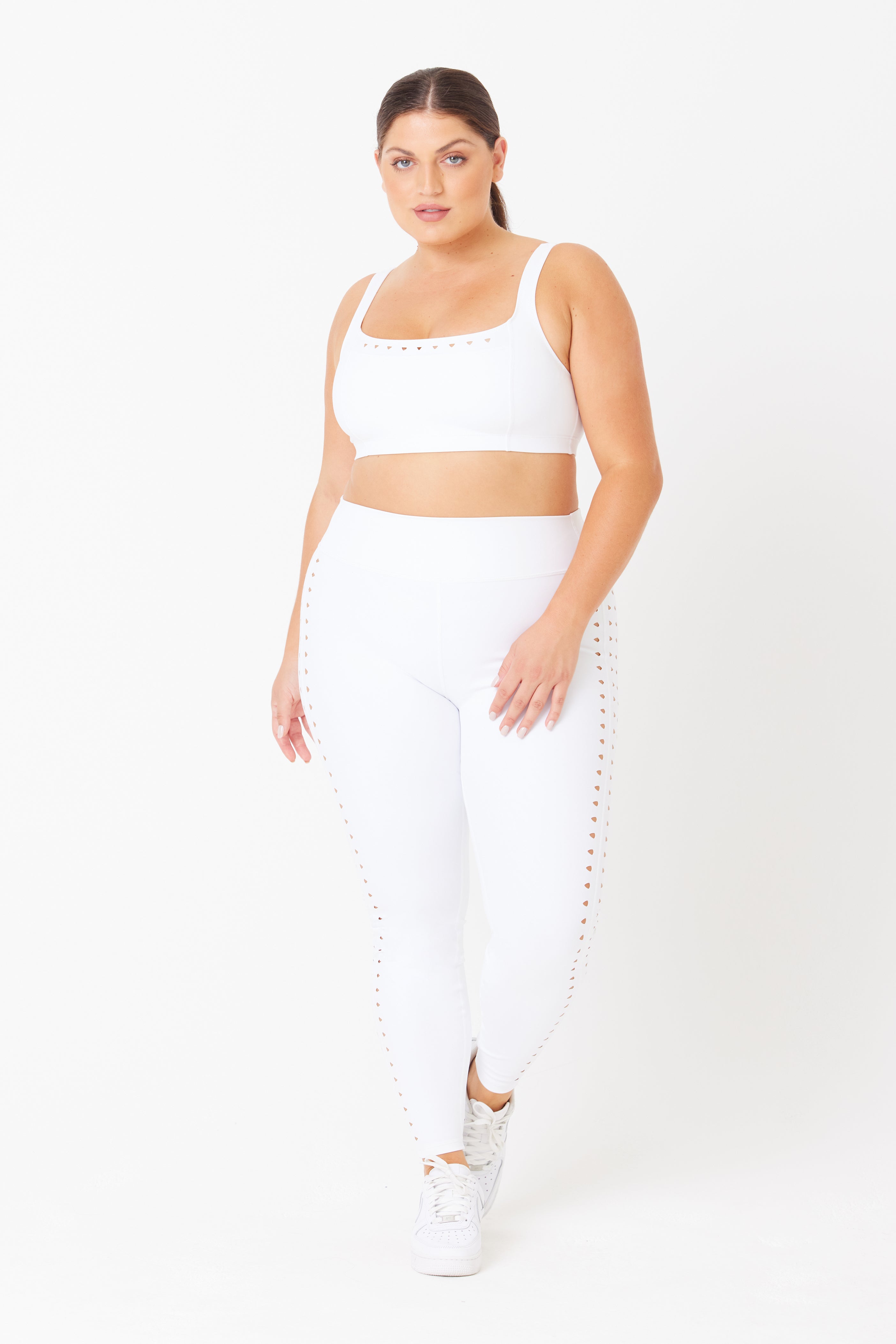 Wearing Gigi C Sports Katrina Legging and matching sports bra, the person stands against a white backdrop. Stylish laser cutouts adorn the sides of both pieces. With white sneakers and tied-back long hair, they perfectly capture the athletic essence of this compression fabric ensemble.