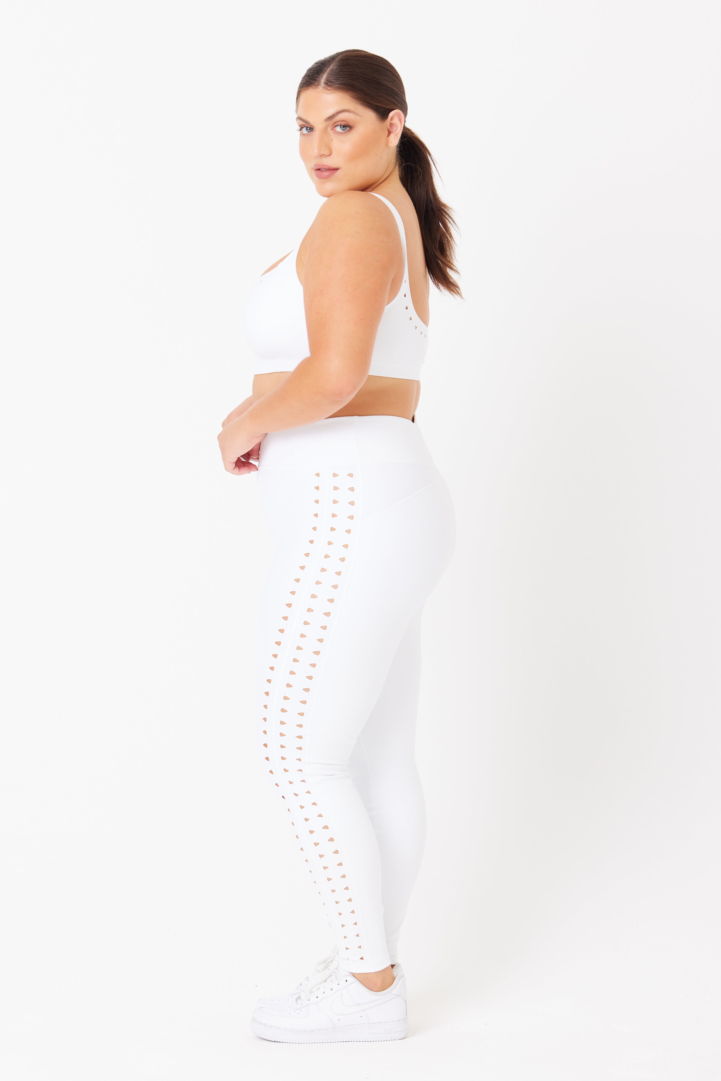 A person with brown hair in a ponytail is wearing a white sports bra and Gigi C Sports Katrina Legging, crafted from compression fabric with stylish laser side cutouts. They pair the look with sleek white sneakers, standing in profile against a pristine white backdrop.