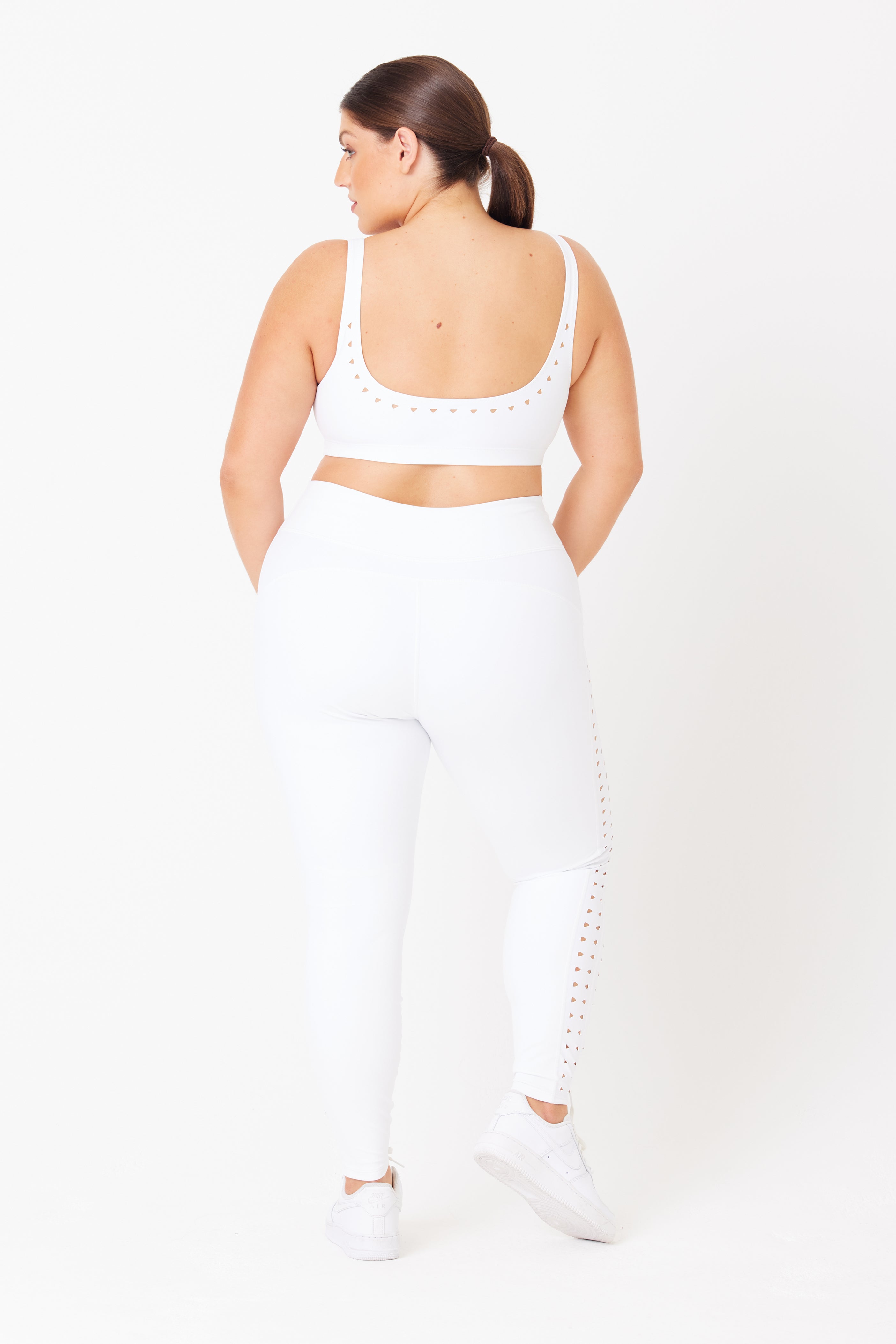 A person with long hair in a ponytail wears a Gigi C Sport white compression set featuring a sports bra and Katrina Legging with laser cutouts. They face away from the camera against a plain white background, paired with pristine white sneakers.