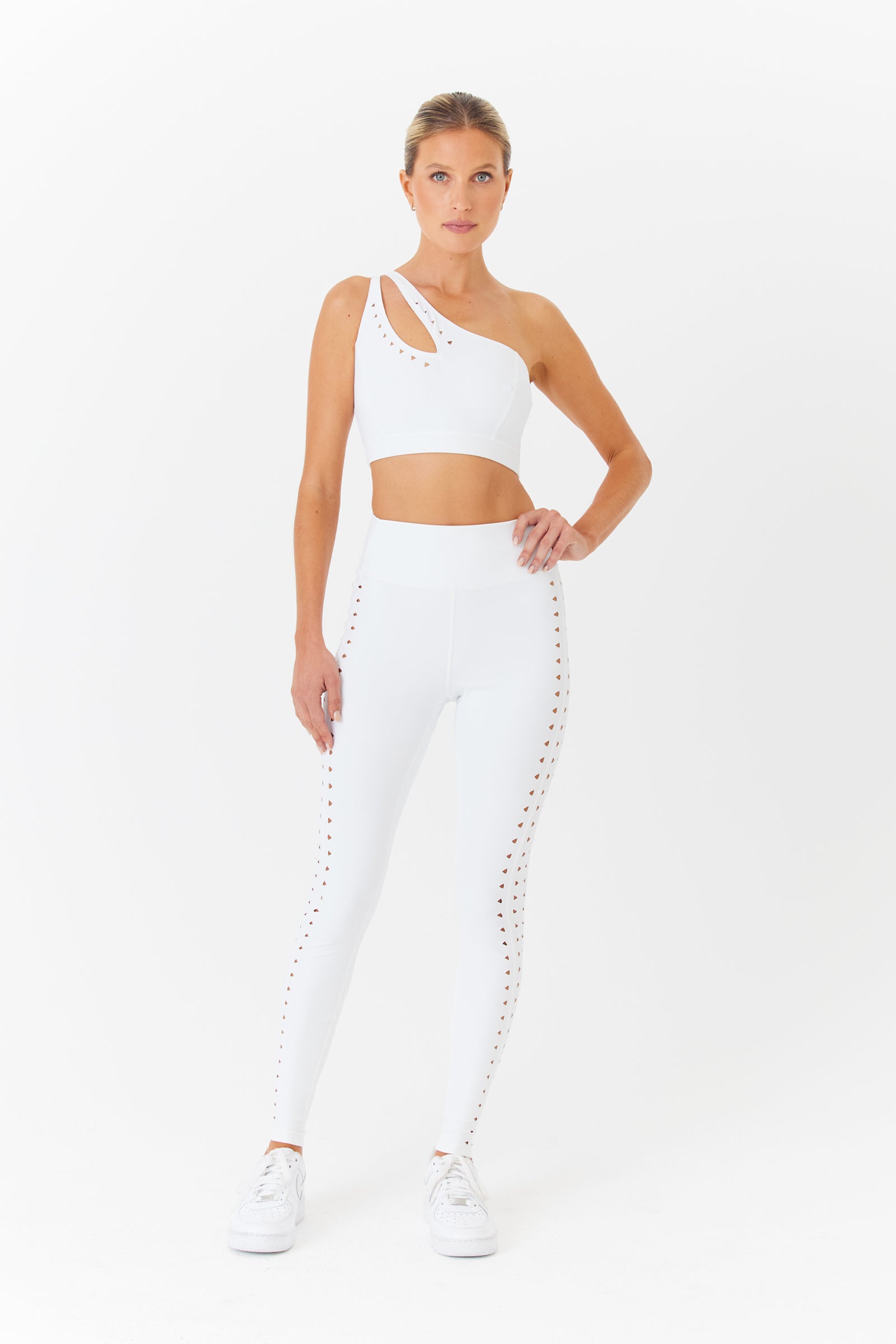 Against a white background, a person models the chic Jacqui Asymmetrical Bra by Gigi C Sport, featuring one-shoulder straps. They pair it with matching high-waisted leggings that have side cut-outs and complete the comfortable, breathable look with white sneakers.