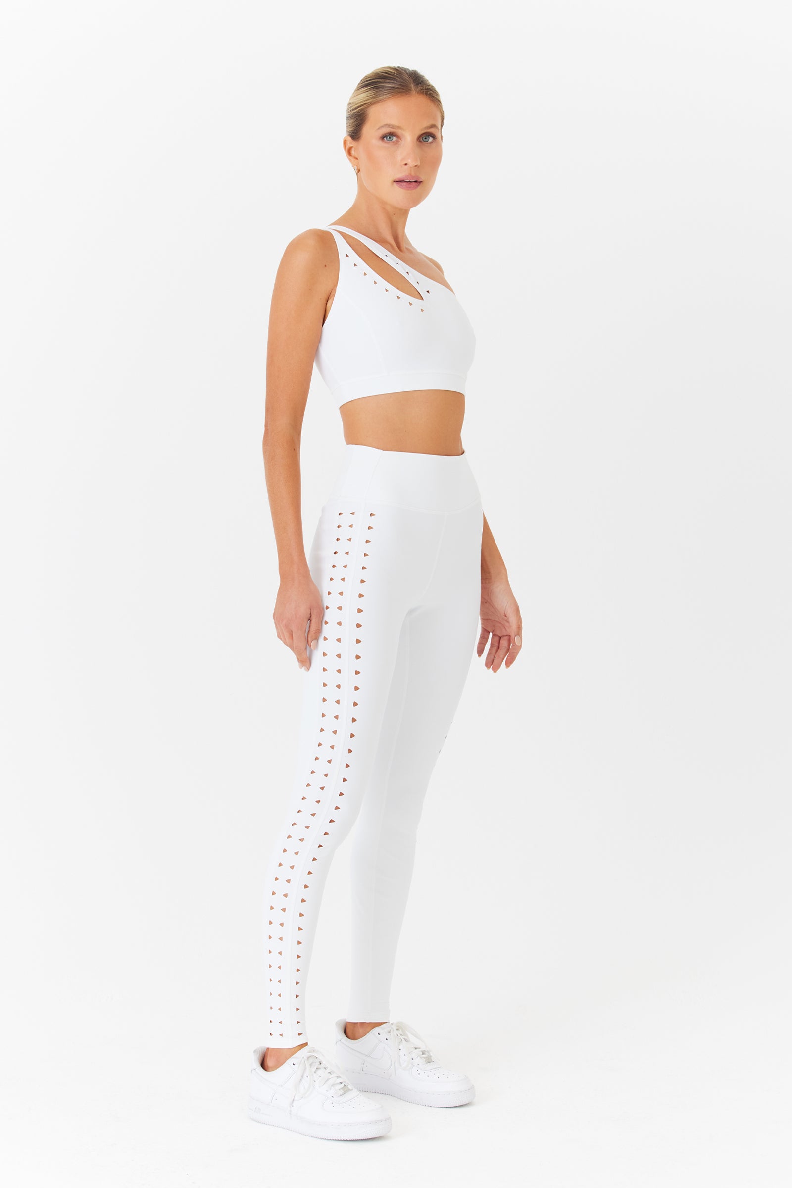 Against a plain white backdrop, a woman models the Gigi C Sport Jacqui Asymmetrical Bra and matching breathable white leggings with cut-out designs. She completes her stylish activewear ensemble with white sneakers.