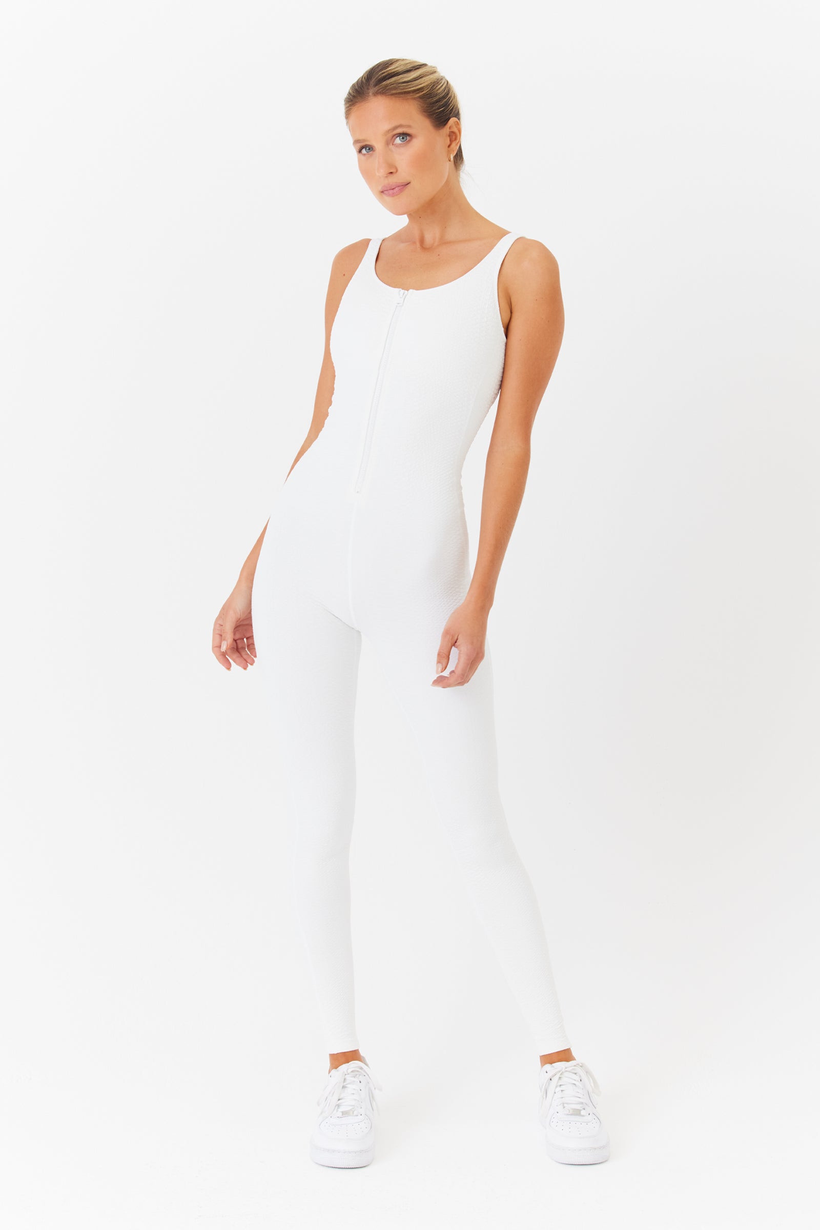 A person poses against a white background in the sleeveless, form-fitting Merit Jumpsuit by Gigi C Sport, made from faux snake jacquard fabric. They wear white sneakers, have one hand on their hip, and stand with a relaxed posture.